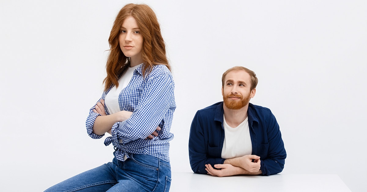 Egy nő komolyan ül az asztal szélén karba tett kézzel, miközben mögötte egy férfi boldogan néz rá, de a nő elérhetetlen, mert nem akar kapcsolatot senkivel.