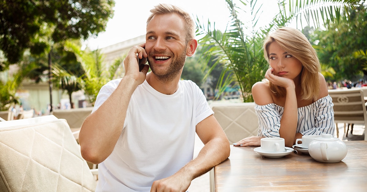 Első randi egy kávézóban, egy férfi és egy nő egymás mellett ül. A férfi épp mosolyogva és elfordulva telefonál, a nő csalódottan és szomorúan néz rá.