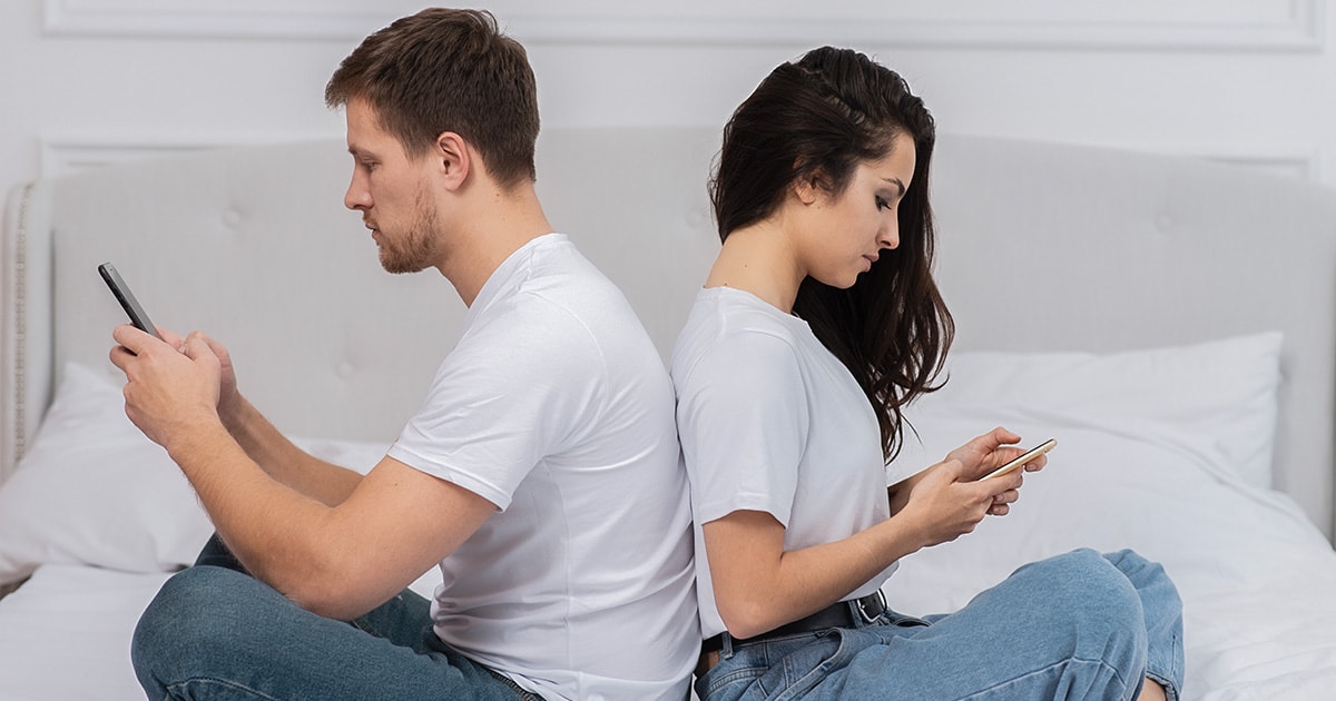 Fiatal pár egymásnak háttal ül az ágyon, mindketten a telefonjukat nézik, érzelmi távolság van köztük.